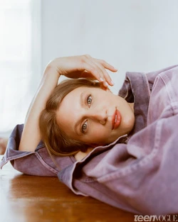 Close-up portrait of a woman in a purple jacket lying on a wooden floor with her hand resting on her head, photographed by Maiwenn Raoult.