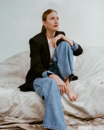 Woman in black blazer and cuffed blue jeans seated on textured fabric backdrop by Maiwenn Raoult