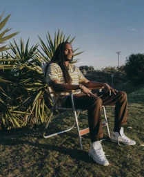 Man with dreadlocks wearing striped polo shirt and brown trousers seated outdoors on a folding chair in Los Angeles by Maiwenn Raoult
