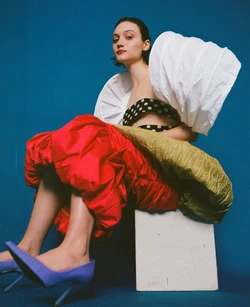 Model seated on a white block wearing an elaborate white puffed sleeve top, polka dot bandeau, red skirt and purple heels against a blue backdrop.