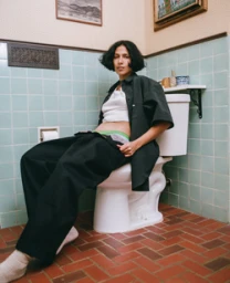 A person with dark bobbed hair wearing a white tank top and black trousers seated on a white toilet in a tiled bathroom.