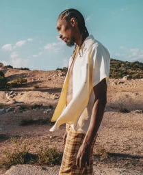 Black man with cornrows wearing a cream button-down shirt and plaid trousers outdoors in a dry landscape