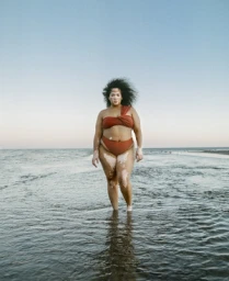 Woman in a rust orange bikini standing in shallow ocean water under a clear sky.