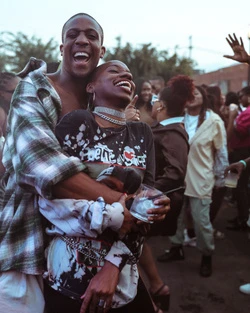 Two smiling Black individuals hugging closely in an outdoor crowd, with a man embracing a woman holding a glass in Los Angeles.