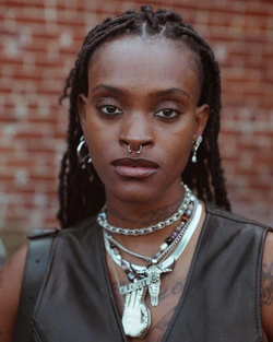 Portrait of a black woman with dreadlocks wearing a septum ring, layered silver necklaces, and a dark vest against a brick wall.