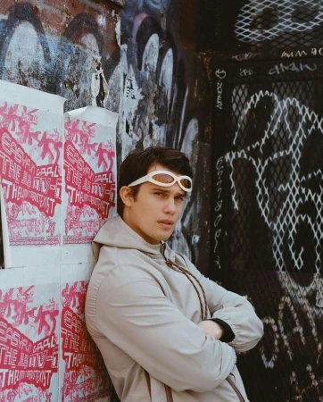 A young man wearing white sunglasses and a beige hoodie leans against a graffiti-covered wall with pasted pink posters.