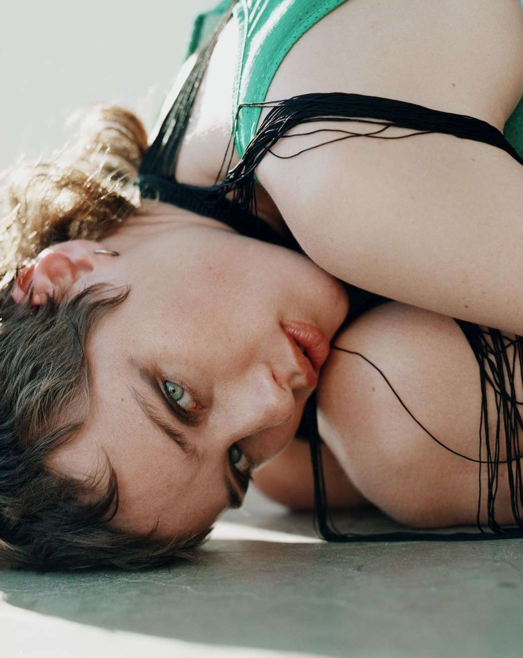 Close-up portrait of a young woman lying down looking at the camera, wearing a green top with black fringe. Indoor, soft lighting.
