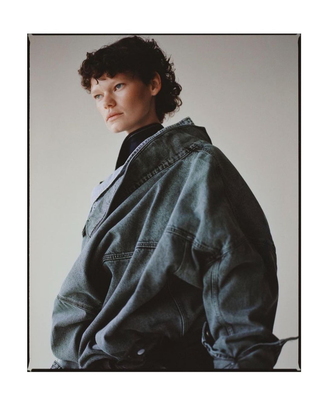 Model with dark curly hair wearing an oversized denim jacket against a neutral studio background by Matthew Priestley.