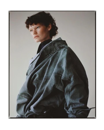 Model with dark curly hair wearing an oversized denim jacket against a neutral studio background by Matthew Priestley.