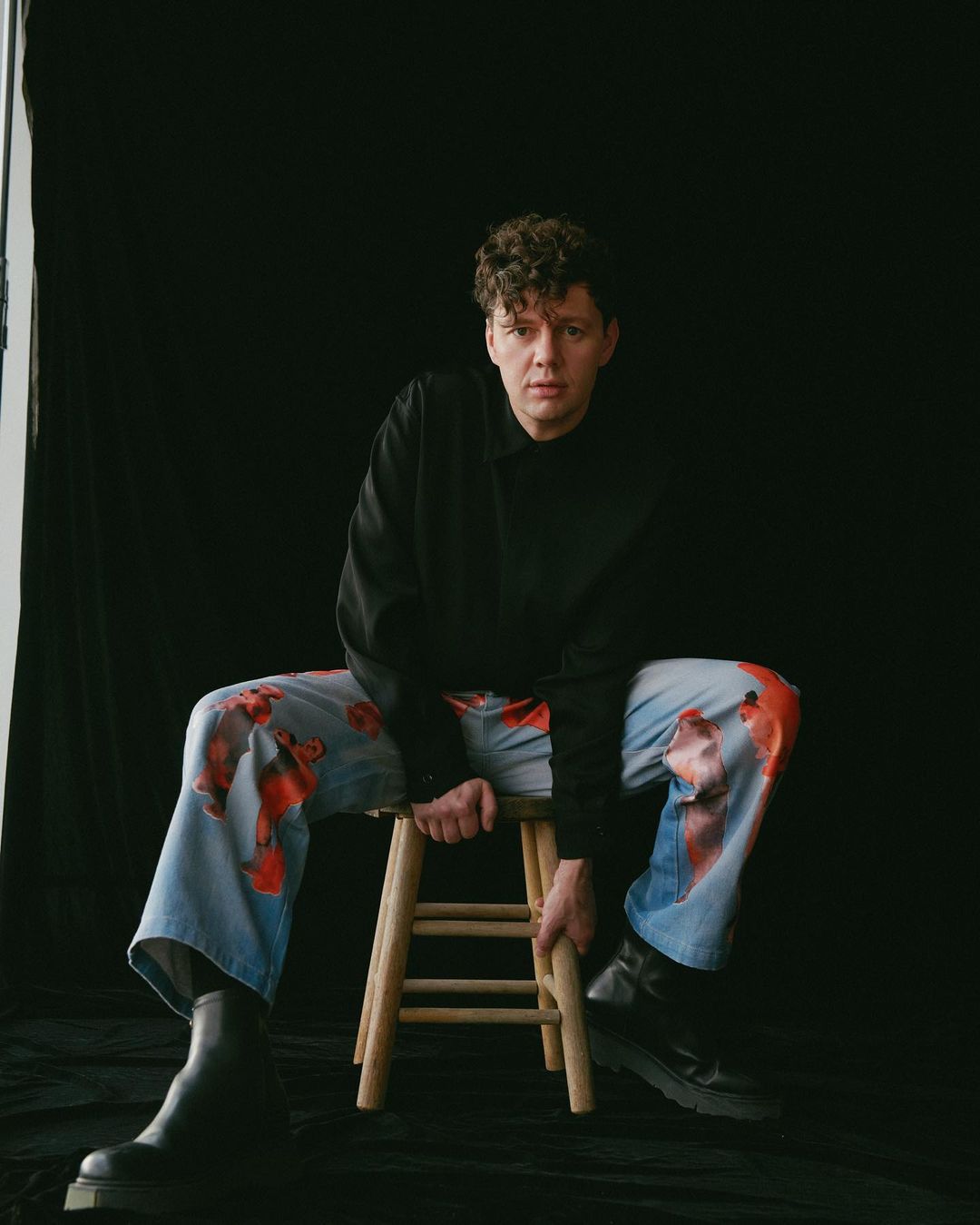 Man with curly hair seated on a wooden stool wearing a black shirt and light blue patterned trousers against a black background.