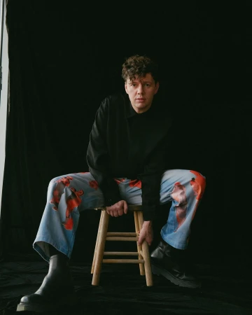 Man with curly hair seated on a wooden stool wearing a black shirt and light blue patterned trousers against a black background.