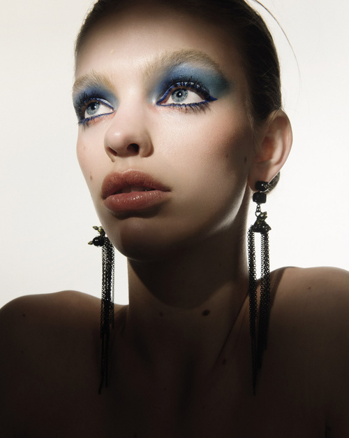 Headshot of a fair-skinned adult woman with vivid blue eyeshadow and long black earrings, looking up in a bright, high-key studio setting.