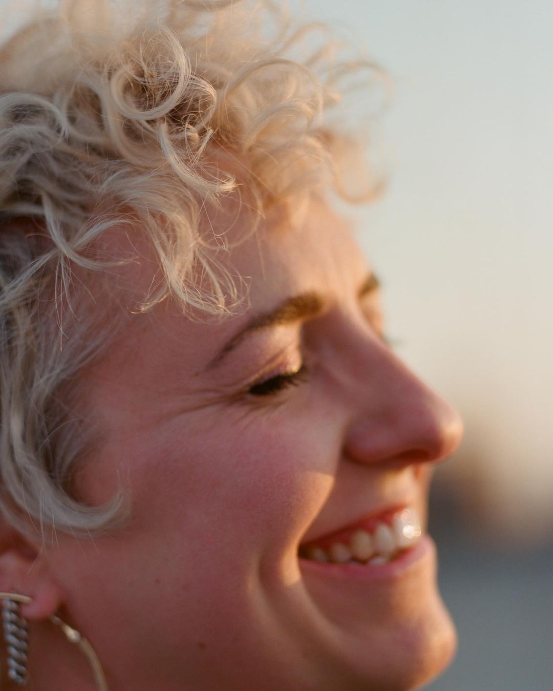 Close-up portrait of a smiling adult female with curly blonde hair. The image captures her face and shoulders in soft, warm, natural light.
