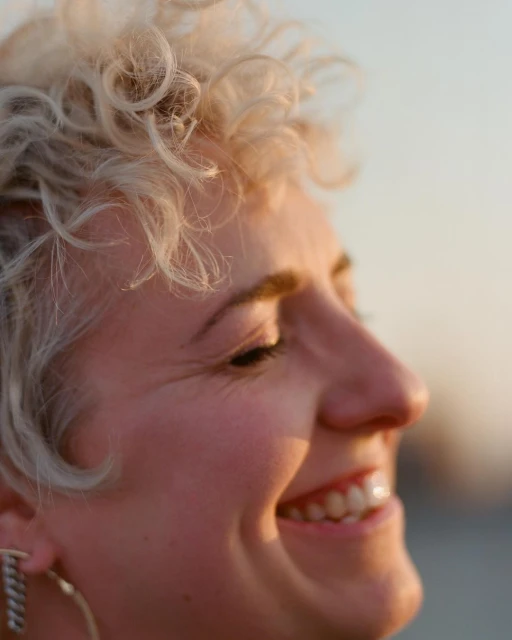 Close-up portrait of a smiling adult female with curly blonde hair. The image captures her face and shoulders in soft, warm, natural light.