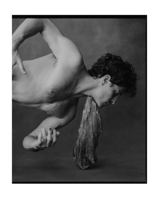 Monochrome portrait of a shirtless adult male figure with curly hair in a bent pose with a piece of wood, set against a dark backdrop.