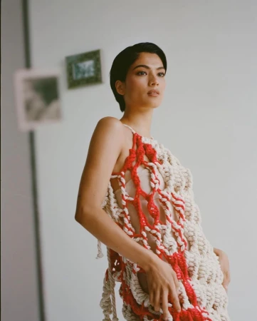 Woman poses wearing red and cream colored crochet dress in studio with neutral background and natural lighting.