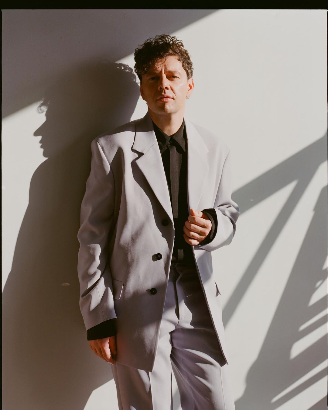 Portrait of an adult male in a light gray suit posing indoors with natural sunlight and a neutral wall background casting a strong shadow.