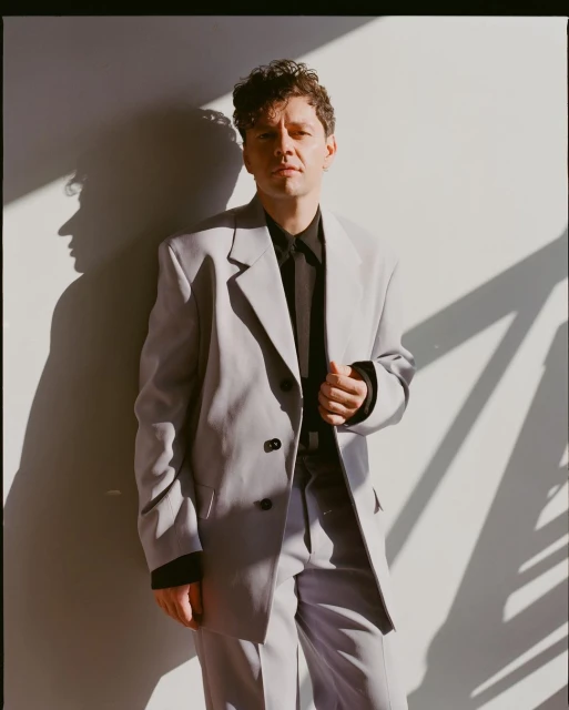 Portrait of an adult male in a light gray suit posing indoors with natural sunlight and a neutral wall background casting a strong shadow.