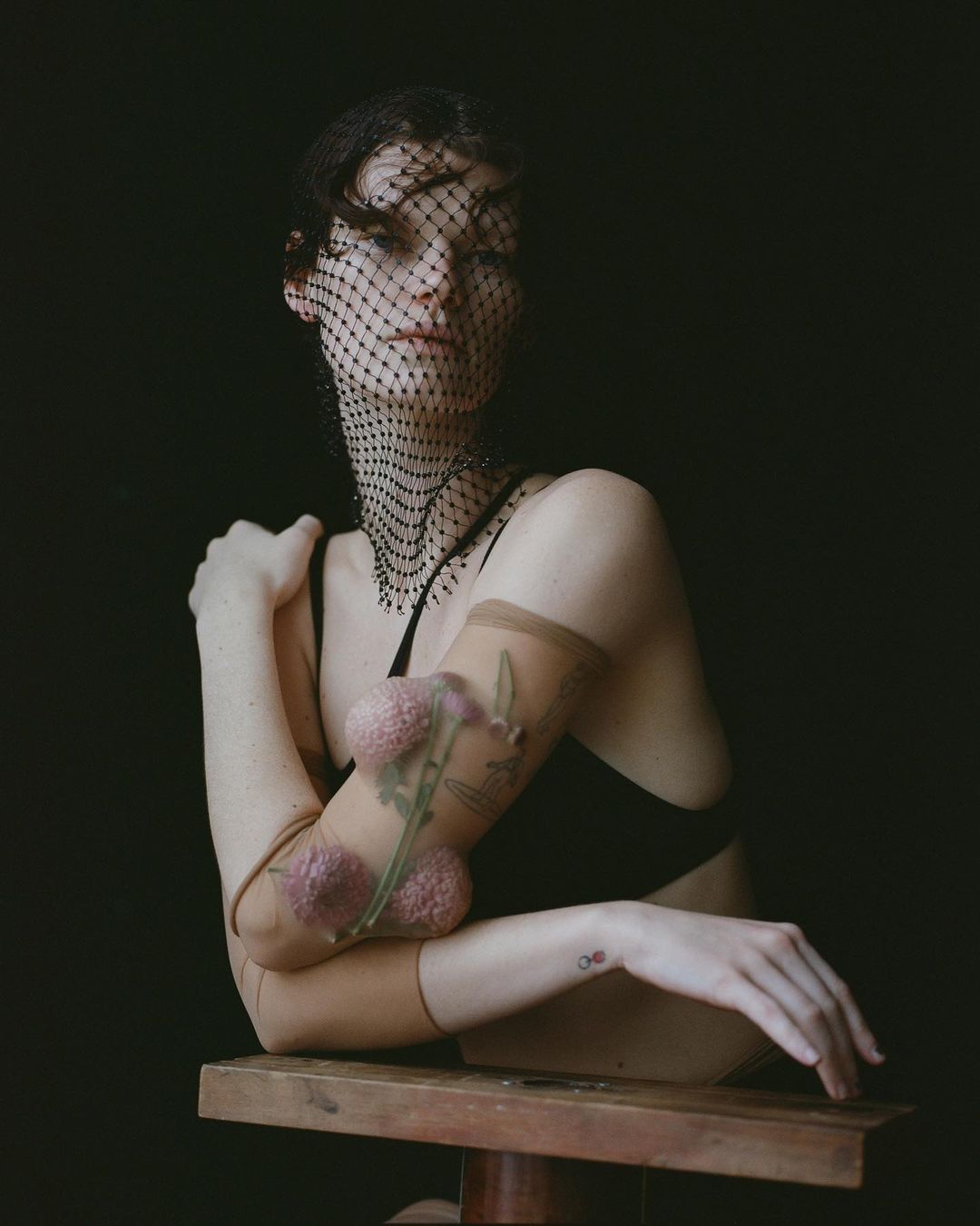 Portrait of a woman in a black top and a mesh head covering, leaning on her arms in front of a dark background in a studio.