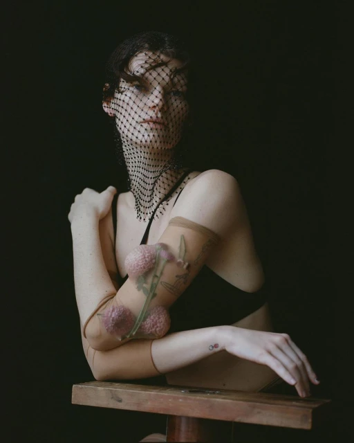 Portrait of a woman in a black top and a mesh head covering, leaning on her arms in front of a dark background in a studio.