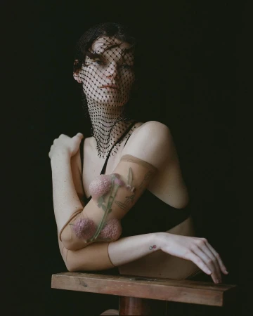 Portrait of a woman in a black top and a mesh head covering, leaning on her arms in front of a dark background in a studio.