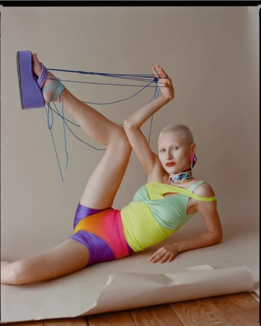 Woman in a neon bodysuit and platform shoe poses on a tan surface in a studio. Strings wrap around her leg and hand.