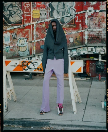 A fashion shot captures a Black model in a stylish gray jacket and lilac pants, standing against a graffiti wall in an urban setting.