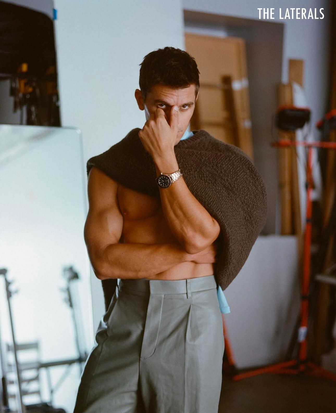 Portrait of adult male in grey trousers and knitwear draped over shoulders, striking a pose in a neutral indoor environment.