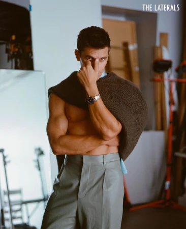 Portrait of adult male in grey trousers and knitwear draped over shoulders, striking a pose in a neutral indoor environment.
