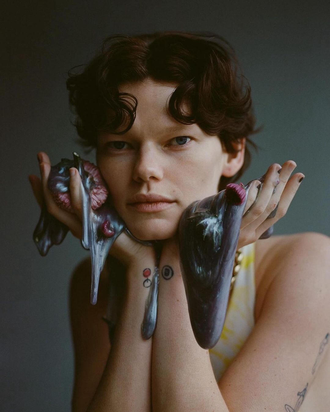 Portrait of an adult with fair skin and short brown hair, holding iridescent liquid with tattoos visible on the forearms against a grey backdrop.