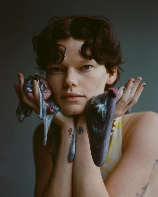 Portrait of an adult with fair skin and short brown hair, holding iridescent liquid with tattoos visible on the forearms against a grey backdrop.