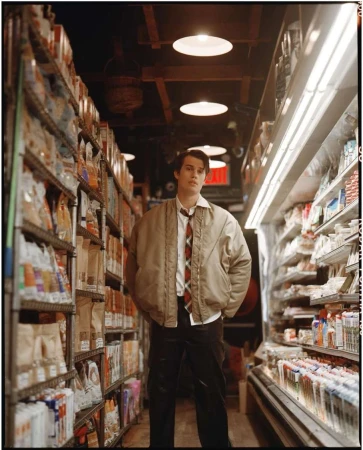 Adult male stands in a grocery store aisle, wearing a khaki jacket and patterned tie with shelves of food products on either side.