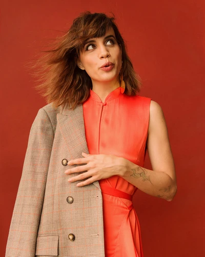 Portrait of a woman in a sleeveless coral dress and plaid jacket, looking upwards against a red backdrop in a studio setting.