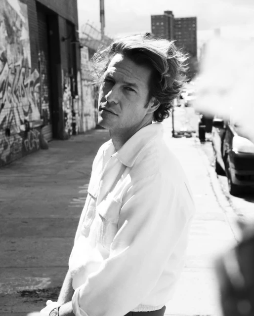Black and white portrait of adult male with wavy hair, wearing a white collared shirt, posing in an urban alleyway with graffiti.