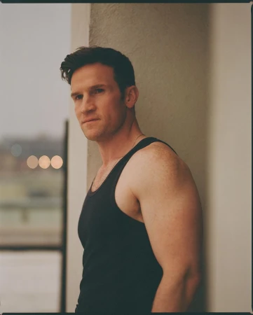 Portrait of a man in a black tank top standing against a neutral concrete pillar with distant city lights blurred in the background