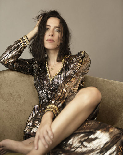Brunette woman in a bronze sequin dress posing on a tan couch in a studio setting, with one hand touching her hair and bare feet visible.