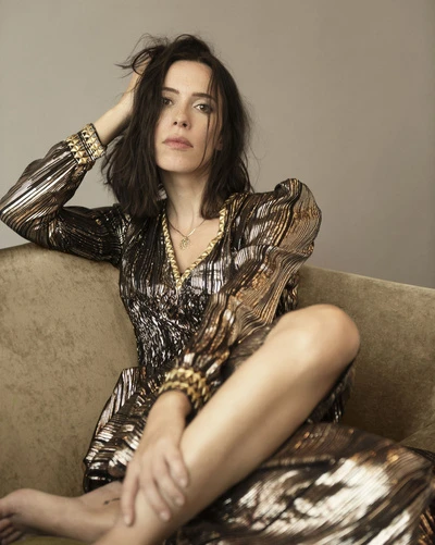 Brunette woman in a bronze sequin dress posing on a tan couch in a studio setting, with one hand touching her hair and bare feet visible.