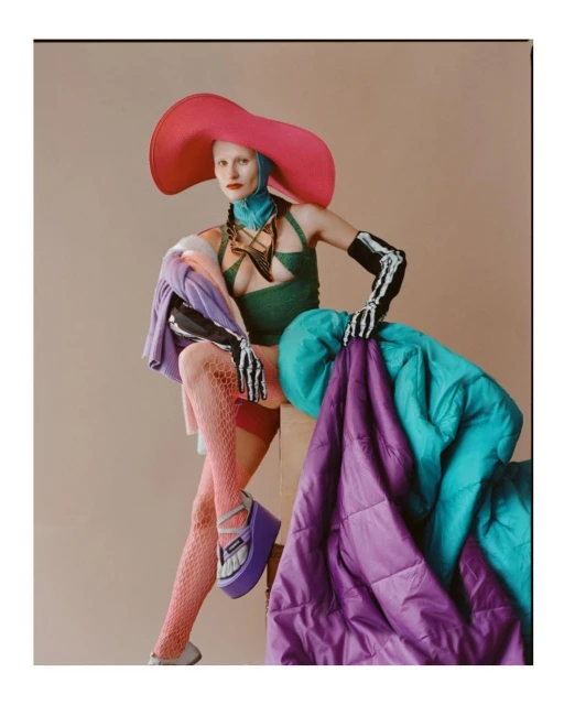 Model wearing an oversized pink hat, green top, fishnet stockings, and platform shoes poses with a colorful quilted blanket.