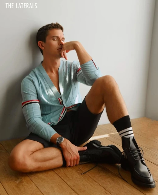 A young adult male model poses indoors, sitting on a wooden floor in a light blue cardigan, black shorts, and black dress boots.