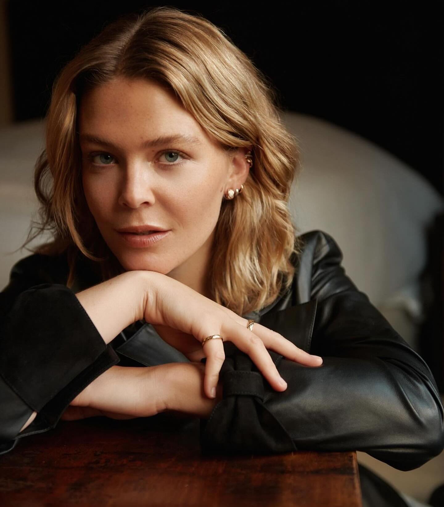 Portrait of a blonde woman in a black leather jacket leaning forward with hands clasped, captured by Matthew Priestley.