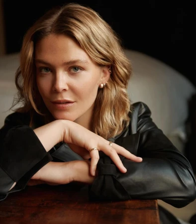 Portrait of a blonde woman in a black leather jacket leaning forward with hands clasped, captured by Matthew Priestley.