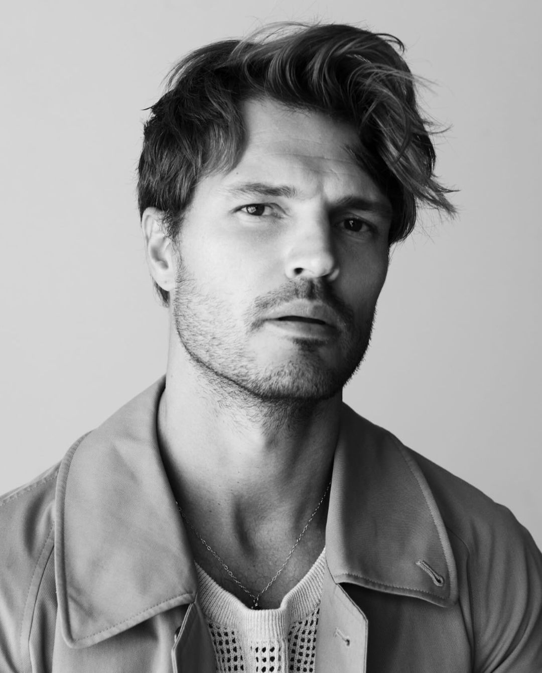Monochrome portrait of an adult male with blond-highlighted hair wearing a coat over a knit shirt. Close-up in a neutral setting.