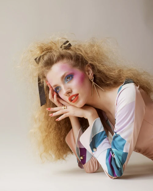 A young woman with bright makeup poses in a studio, wearing a patterned top and a hair ribbon. Her blonde hair is styled in a wavy fashion.
