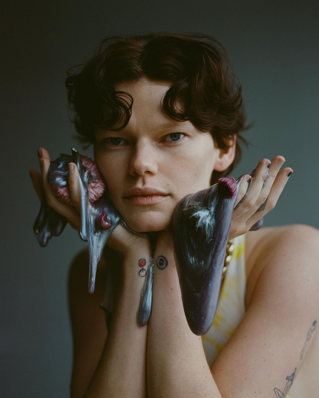 Portrait of a person with short, dark, curly hair holding glossy, purple sculptural objects with hands covered in iridescent paint.