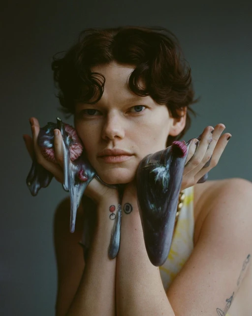Portrait of a person with short, dark, curly hair holding glossy, purple sculptural objects with hands covered in iridescent paint.