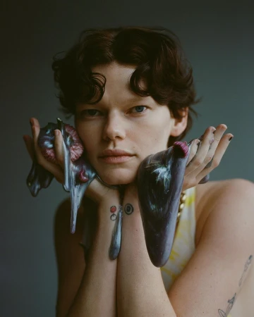 Portrait of a person with short, dark, curly hair holding glossy, purple sculptural objects with hands covered in iridescent paint.