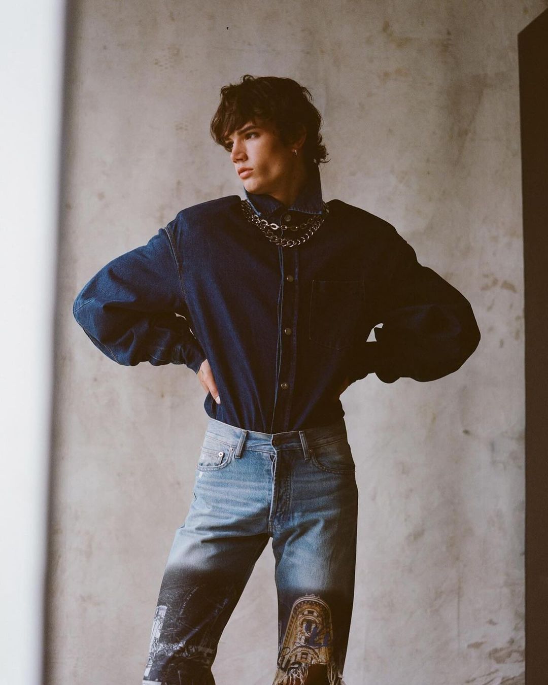 Studio shot of a young adult man in denim shirt and printed denim pants with chains, hands on hips, posing in front of grey wall.