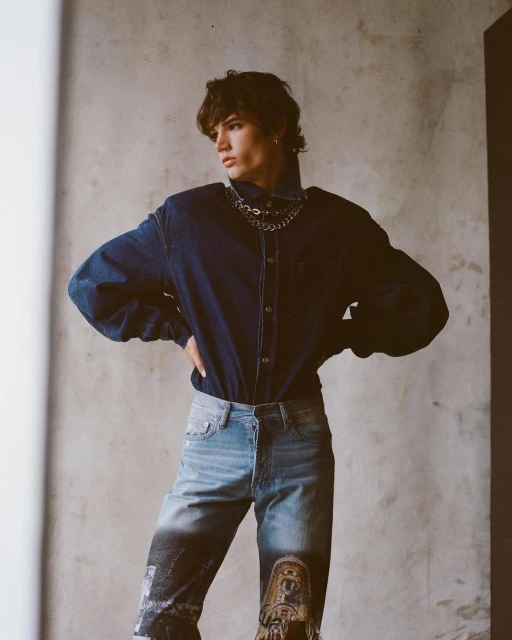 Studio shot of a young adult man in denim shirt and printed denim pants with chains, hands on hips, posing in front of grey wall.