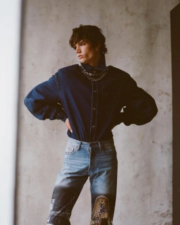 Studio shot of a young adult man in denim shirt and printed denim pants with chains, hands on hips, posing in front of grey wall.