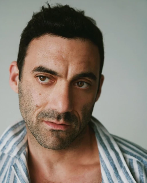 Portrait of an adult male with dark hair and light eyes, wearing a striped shirt, looking off to the side against a neutral background.